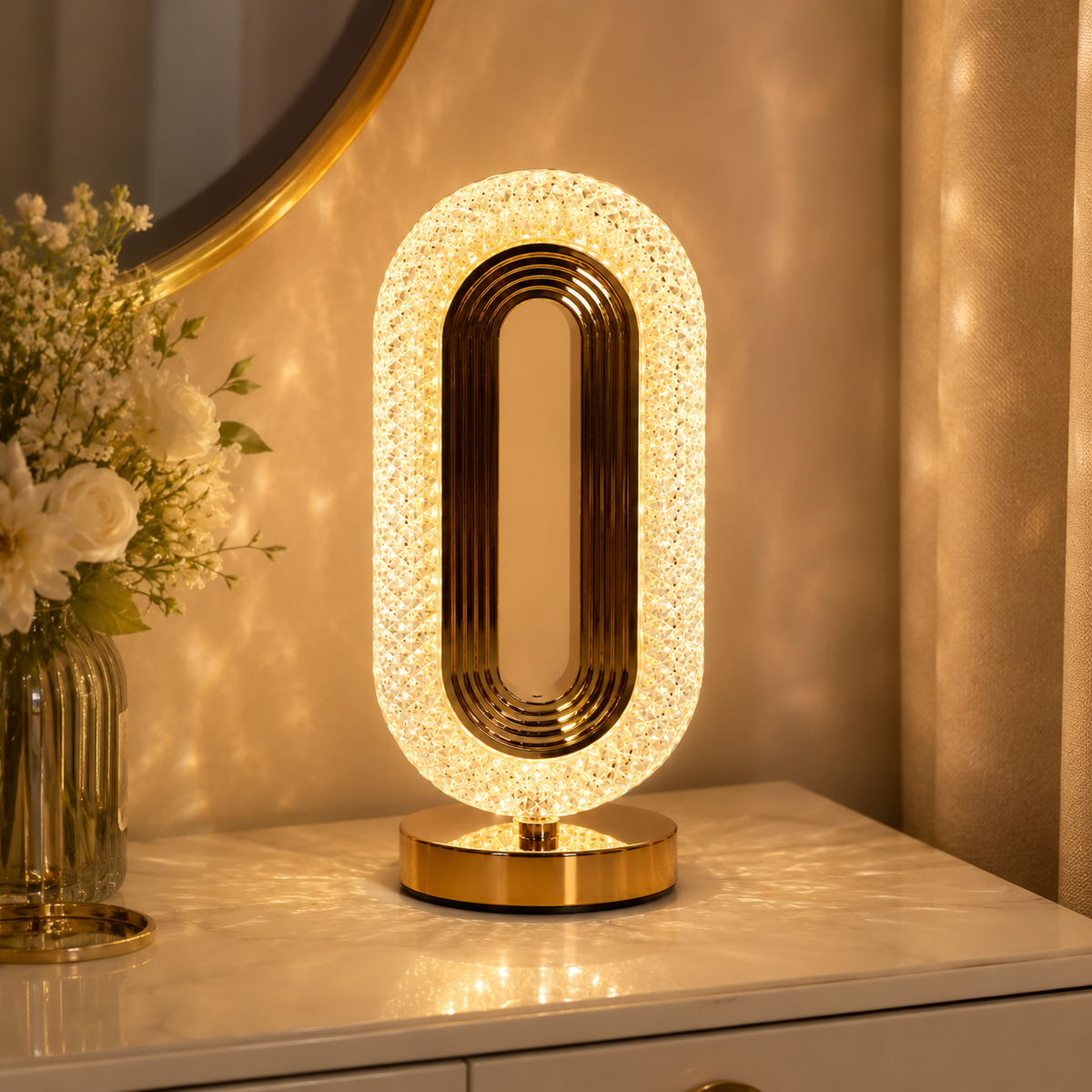 Gold oval crystal table lamp on a marble dresser beside a mirror and flowers